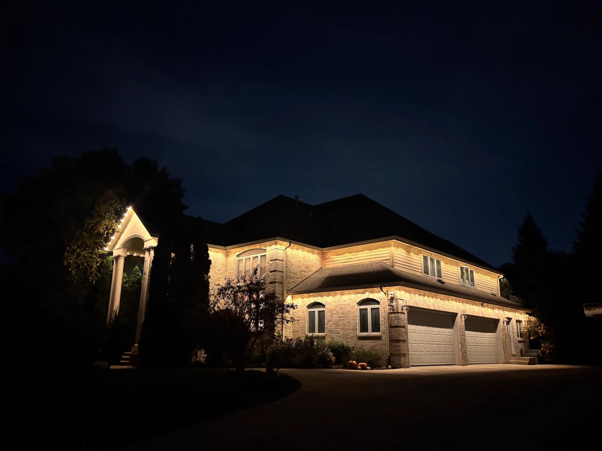Real Kussmaul Co. permanent lighting in warm white along a brick two-story home, its columned front portico and three-car garage at night