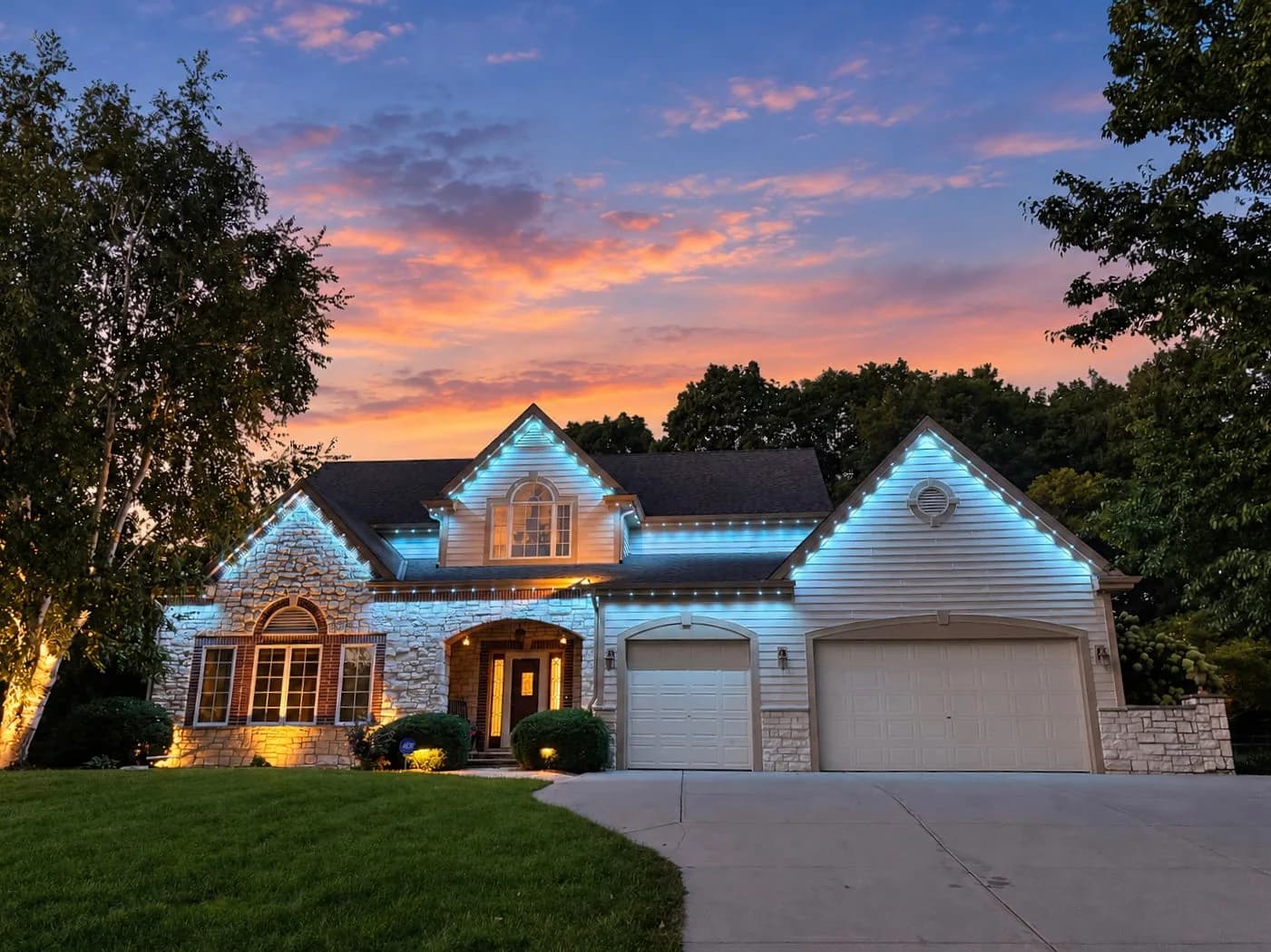 Real Kussmaul Co. permanent lighting in crisp cool white across a stone two-story home at sunset